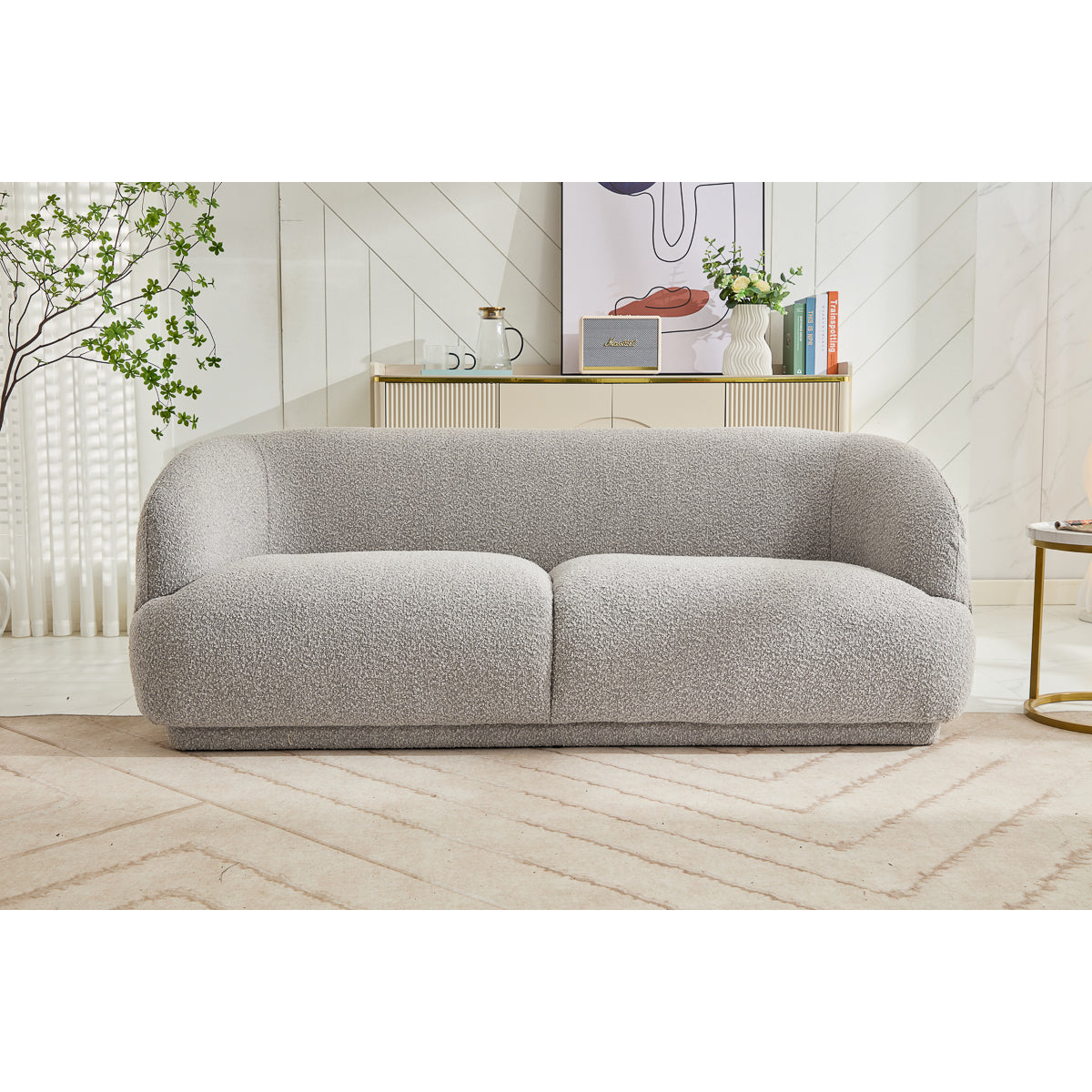 A modern living room showcases the Avriio Madrid 3 Seater Taupe Sofa on a beige patterned rug, paired with a gold console table, decor accents, lush green plants, and convenient Home Delivery options.