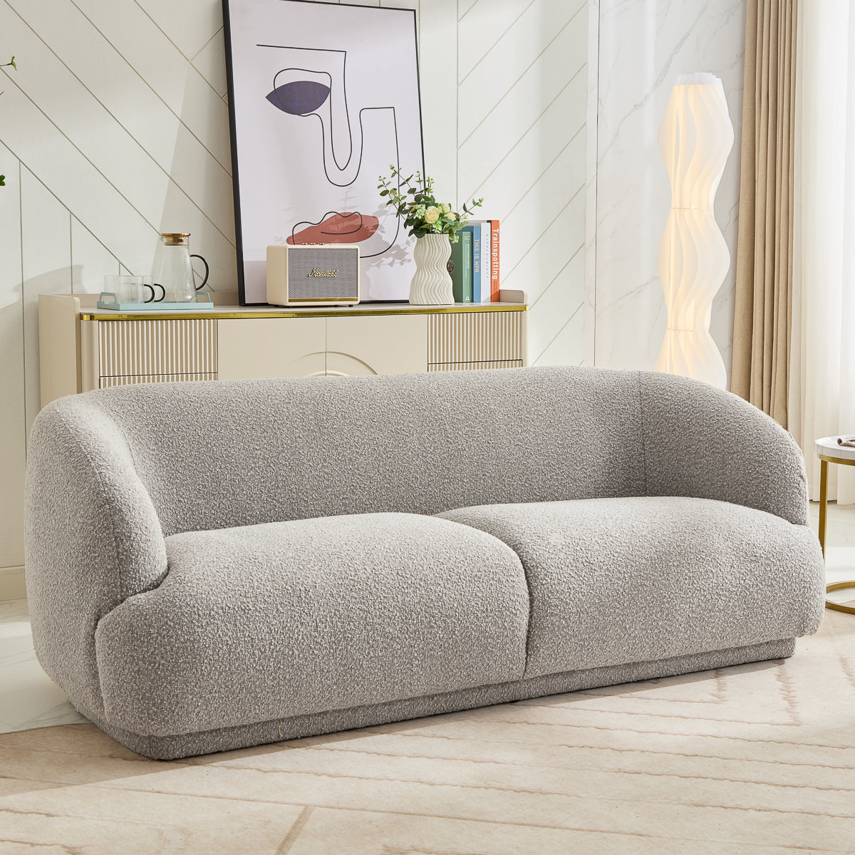 A modern living room features the Avriio Madrid 3 Seater Taupe Sofa with a curved, textured design. A sideboard holds books, vases, and abstract art. Enjoy convenient home delivery and White Glove setup options for easy installation.