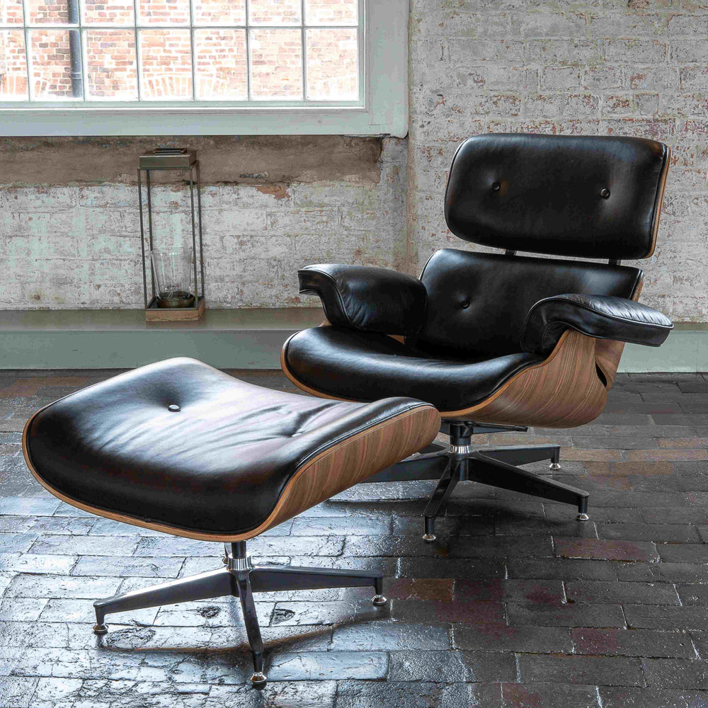 The Carlton Furniture Eames Style Lounge Chair and Stool, featuring leather upholstery and a walnut finish, is placed on a dark tiled floor by a large window against a textured white brick wall.