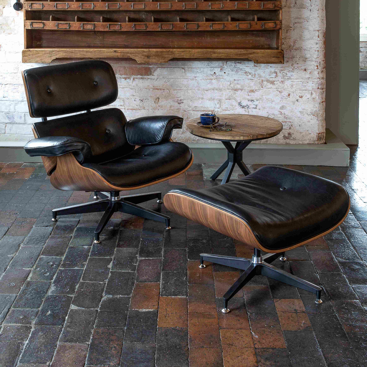 
                  
                    A Carlton Furniture Eames Style Lounge Chair and Stool with black leather upholstery sits next to a small round wooden table holding a coffee cup, set on a rustic brick floor against a brick wall with wooden shelves.
                  
                