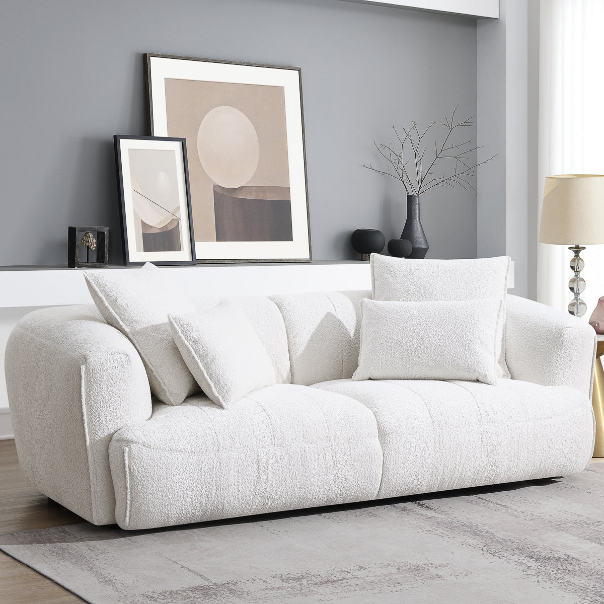 A modern living room features the Avriio Kastoria Short Leg 3 Seater White Silver Sofa with textured fabric, four white cushions, abstract art on a shelf, and a black vase with branches. Enjoy White Glove Home Delivery to your room of choice.