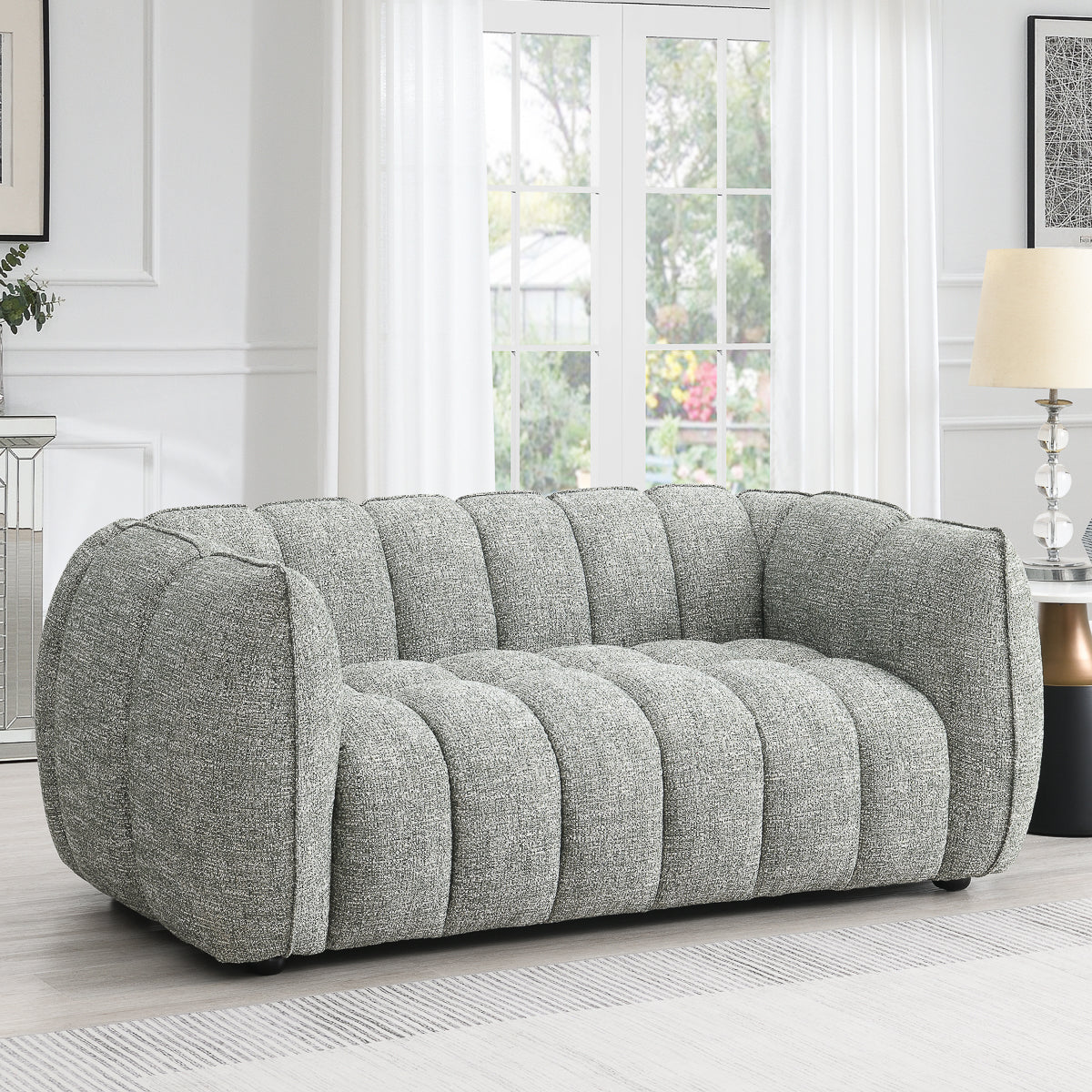The Avriio Vienna 2 Seater Winter Moss Fluted Fabric Sofa adds modern style to a bright living room with white walls, sheer curtains, a lamp, and a large window overlooking a lush garden. Enjoy hassle-free White Glove Home Delivery.